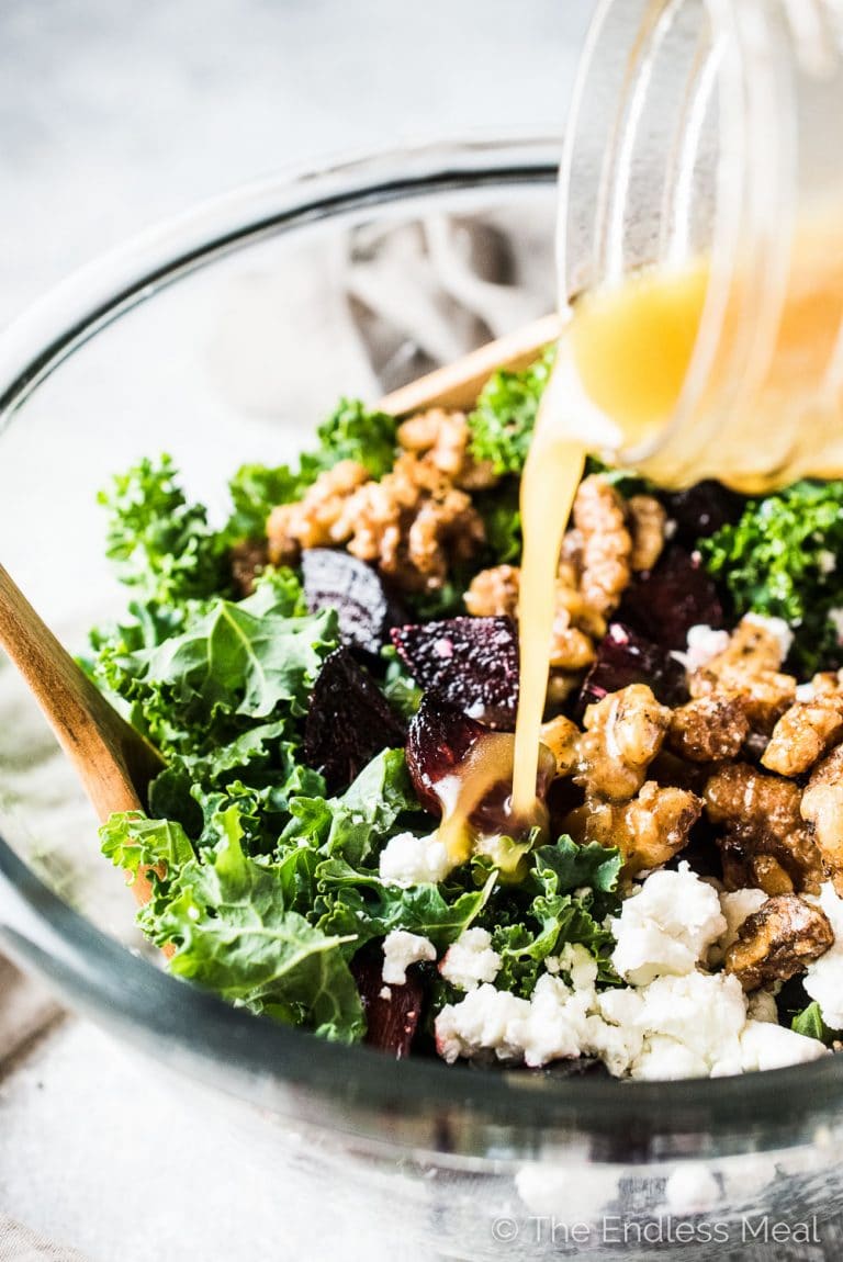 A glass bowl of roasted beet and kale salad with dressing being poured on it.