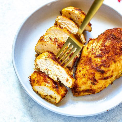 A close-up of two baked chicken breasts on a white plate, one sliced and one left whole, with a fork picking up a sliced piece.