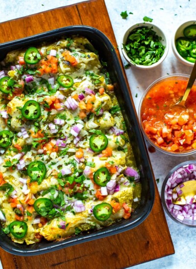 Chicken enchiladas verdes in a black casserole dish next to bowls of cilantro, sliced jalapeno, salsa, and diced red onion.