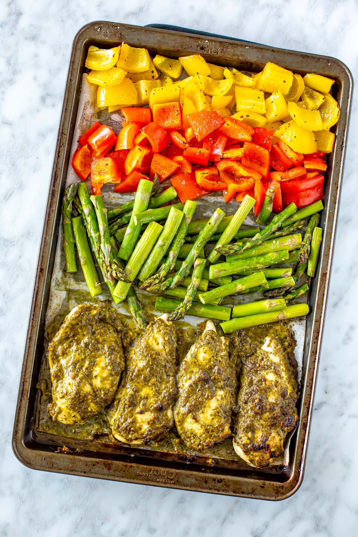 A sheet pan of pesto coated chicken cutlets with chopped asparagus, red pepper and yellow pepper.