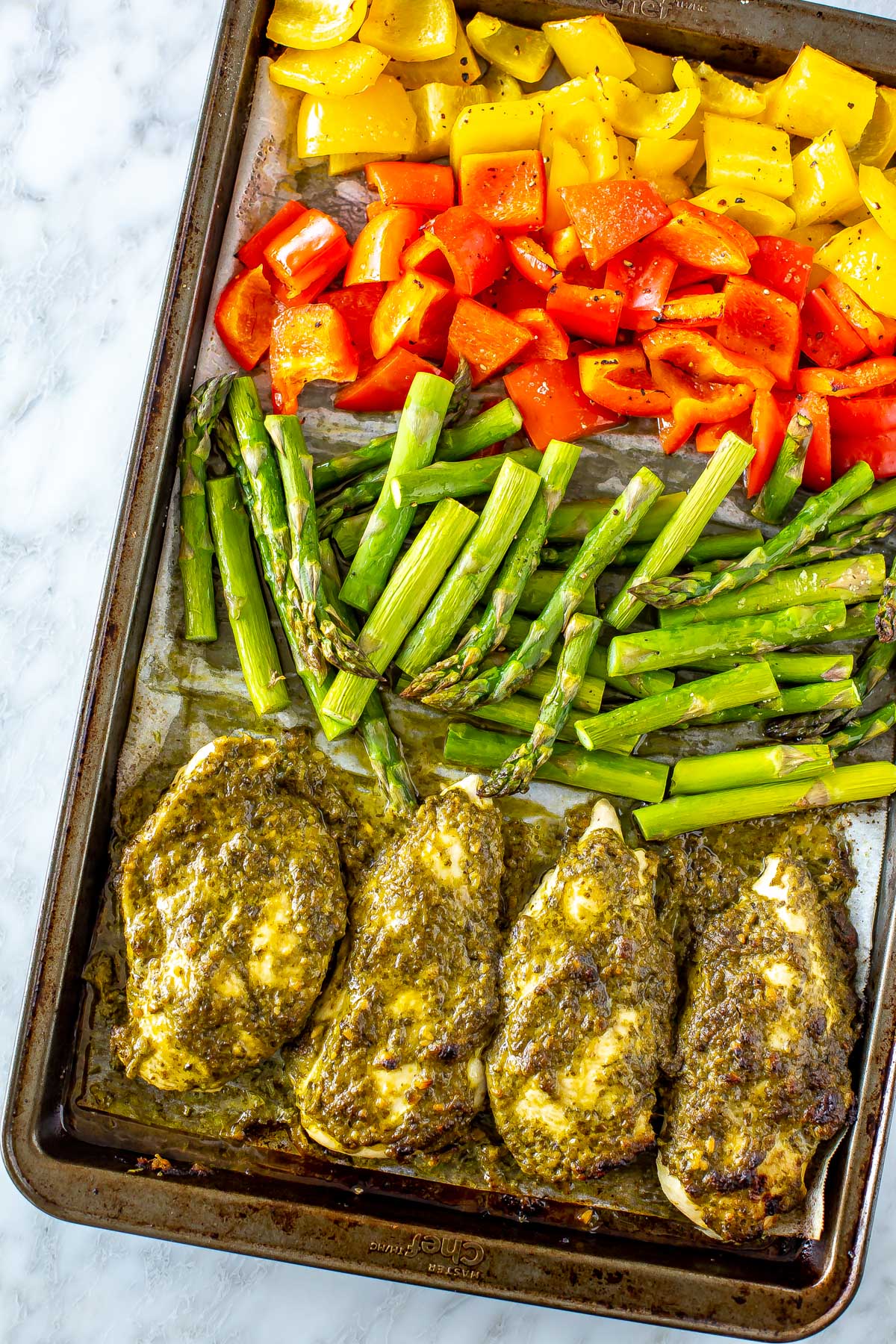 A sheet pan of pesto coated chicken cutlets with chopped asparagus, red pepper and yellow pepper, zoomed in on an angle.