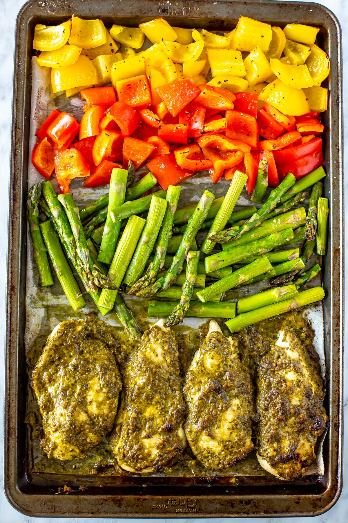A sheet pan of pesto coated chicken cutlets with chopped asparagus, red pepper and yellow pepper, close up.
