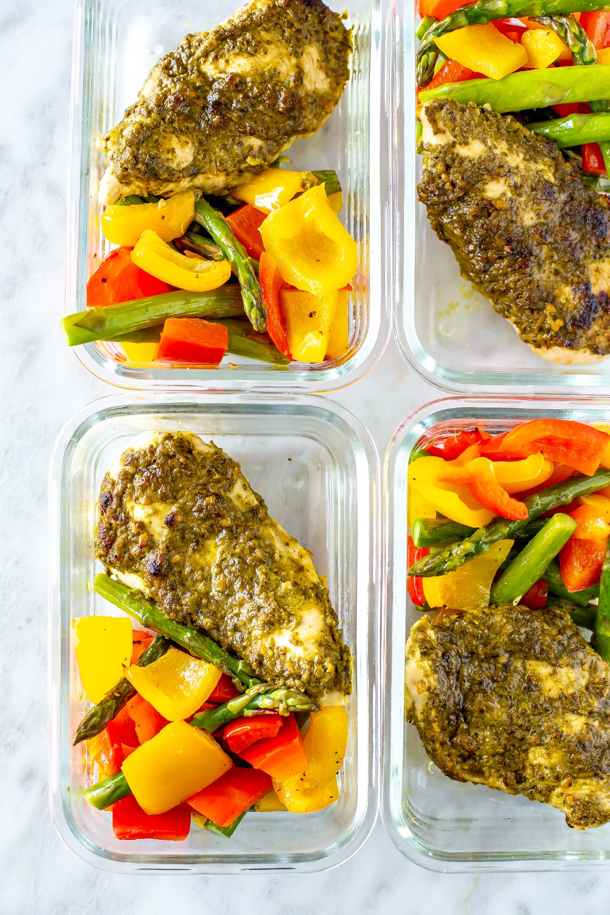Pesto coated chicken cutlets with asparagus, red pepper and yellow pepper in square glass meal prep containers, zoomed in on the bottom left hand corner.