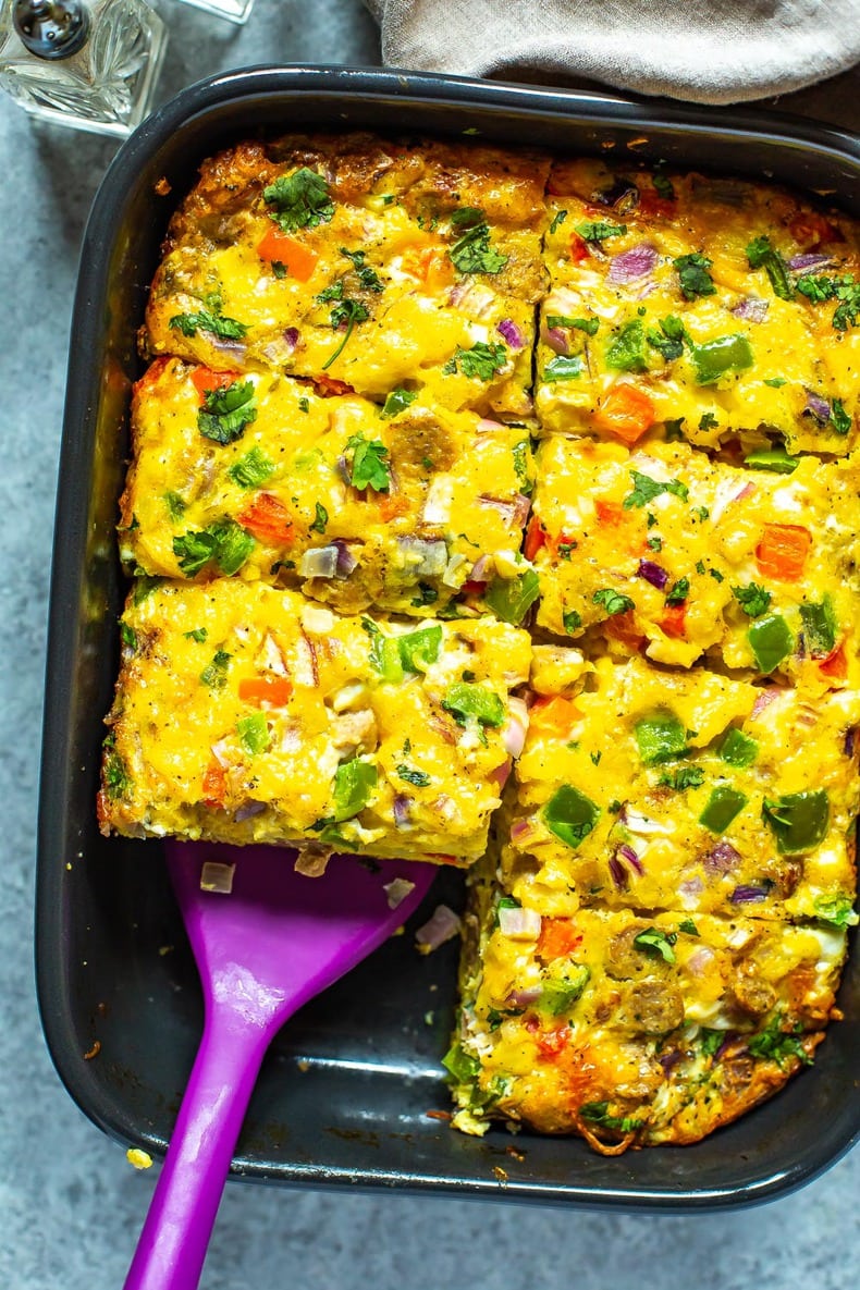 A cooked sausage hashbrown breakfast casserole in a black casserole dish cut into 8 pieces with a spatula picking up a piece.