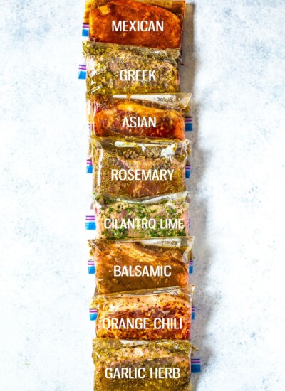 8 different freezer bags stacked on top of each other, each containing a different kind of marinated steak, labelled "Mexican", "Greek", "Asian", "Rosemary", "Cilantro Lime", "Balsamic", "Orange Chili" and "Garlic Herb".