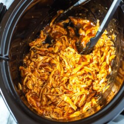 A close-up shot of a slow cooker filled with shredded BBQ chicken with open tongs inside.