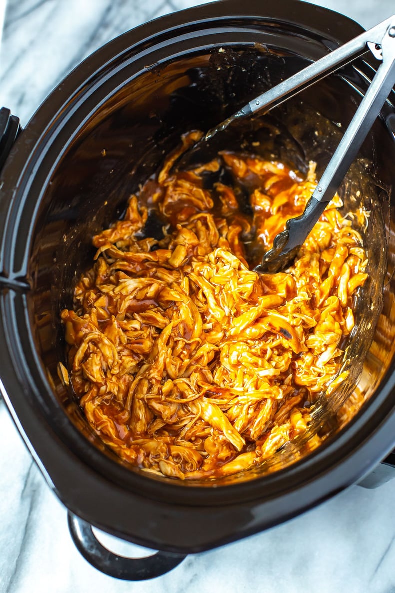 A close-up shot of a slow cooker filled with shredded BBQ chicken with open tongs inside.