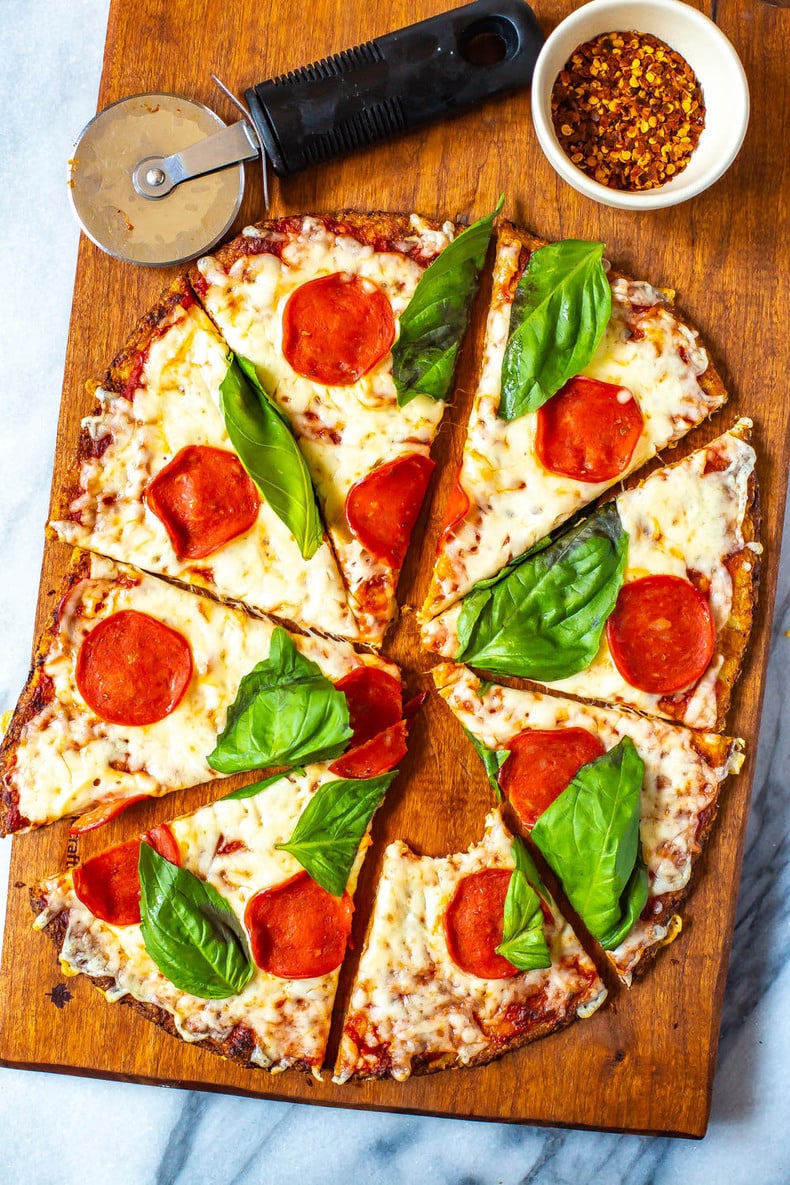 Cauliflower Crust Pizza with pepperoni and basil, cut into slices on a cutting board. One slice has a bite taken out of it.