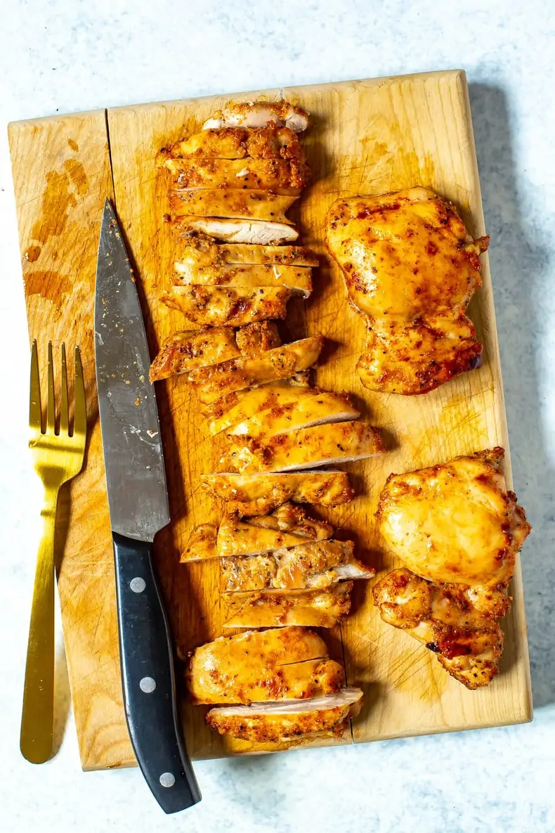 A cutting board pictured with a fork, knife, and chicken thighs being sliced.