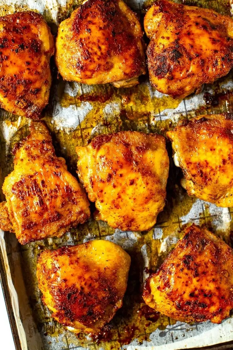 A close-up of seasoned baked chicken thighs on a sheet pan with prepared with parchment paper.