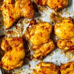 A close-up of seasoned baked chicken thighs on a sheet pan with prepared with parchment paper.