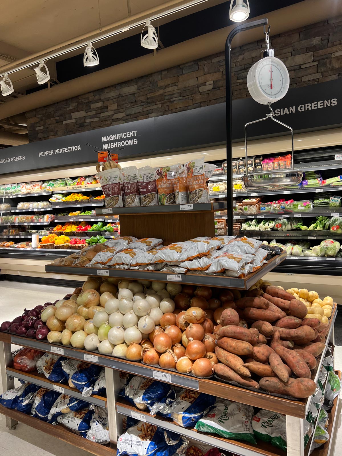 The produce aisle of a grocery store with sweet potatoes and onions in the middle.