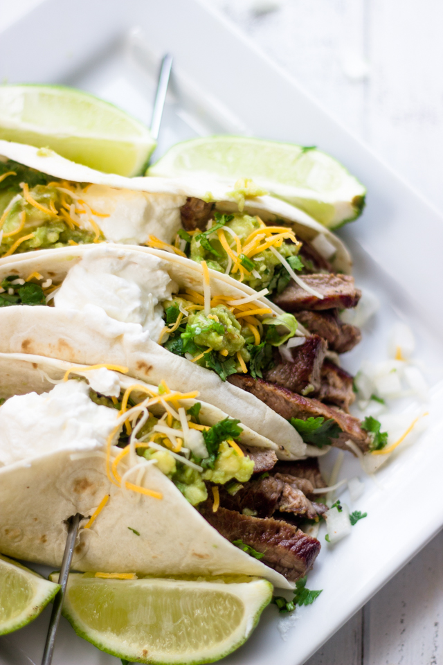 Three Mexican steak tacos side by side topped with guacamole, cilantro, cheese and sour cream.