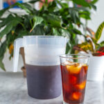 A pitcher of cold brew coffee next to a class of black cold brew coffee with ice.