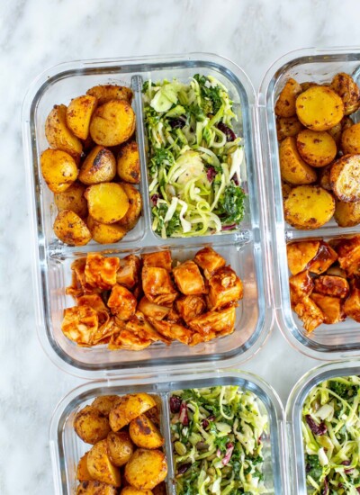 A close-up of a meal prep container with roasted baby potatoes, kale salad and baked BBQ chicken inside.