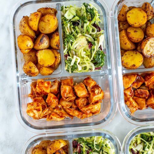 A close-up of a meal prep container with roasted baby potatoes, kale salad and baked BBQ chicken inside.