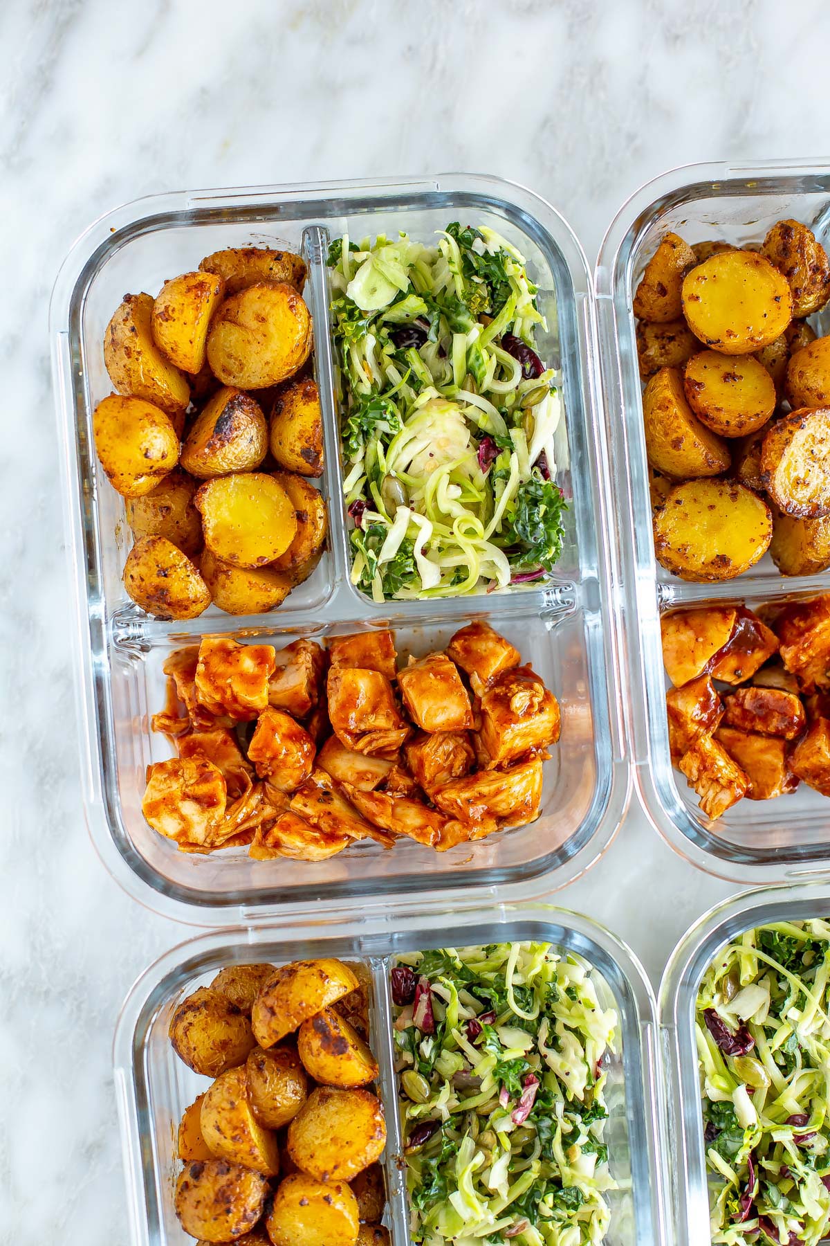 A close-up of a meal prep container with roasted baby potatoes, kale salad and baked BBQ chicken inside.