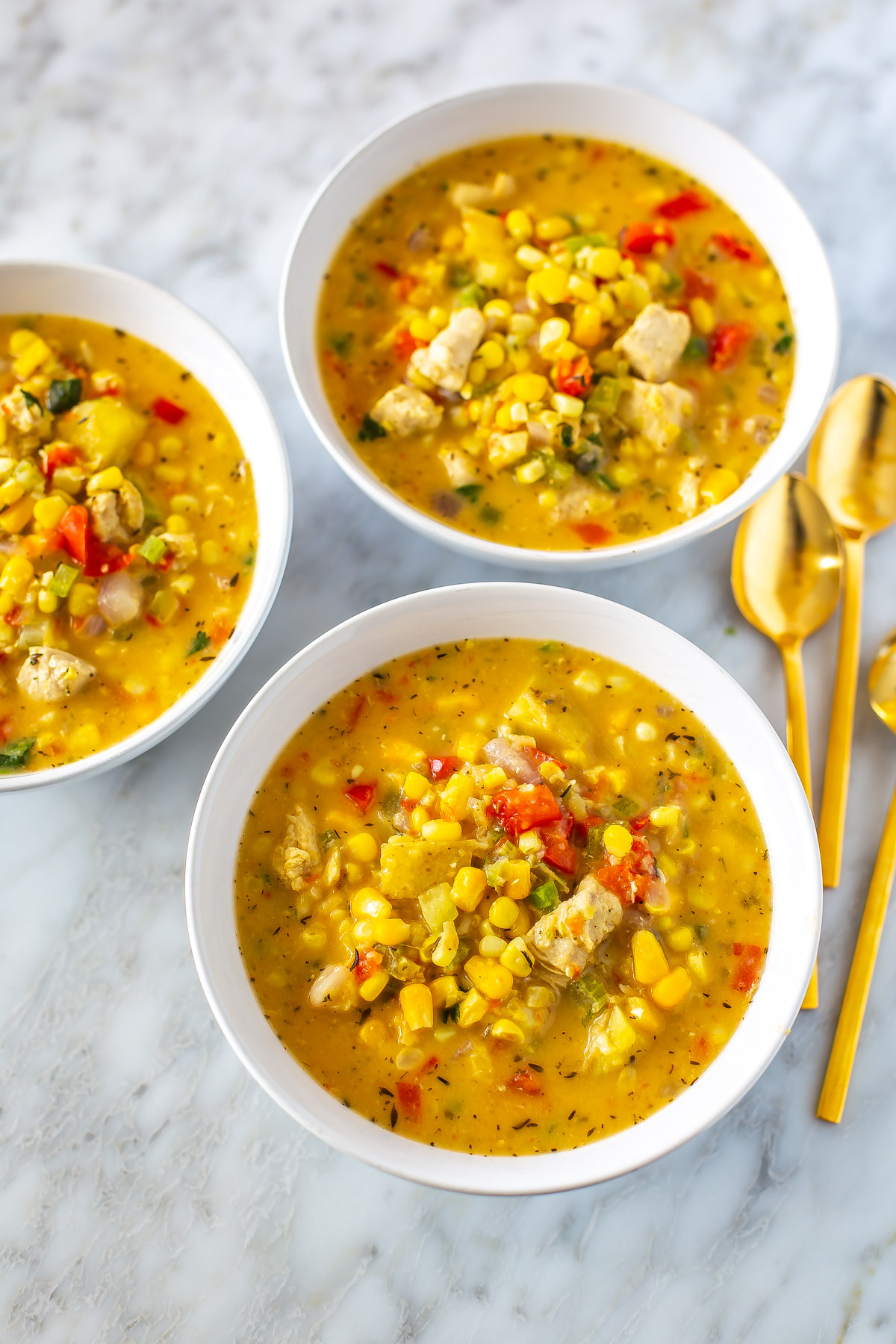 Three bowls of chicken corn chowder with three gold spoons placed next to them.