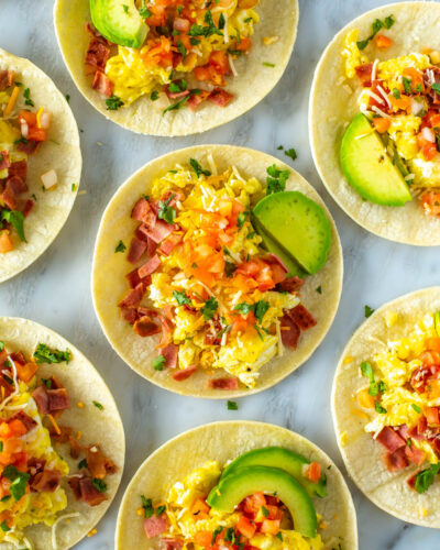 An overhead shot of breakfast tacos topped with eggs, bacon, pice de gallo, avocado and cheese.