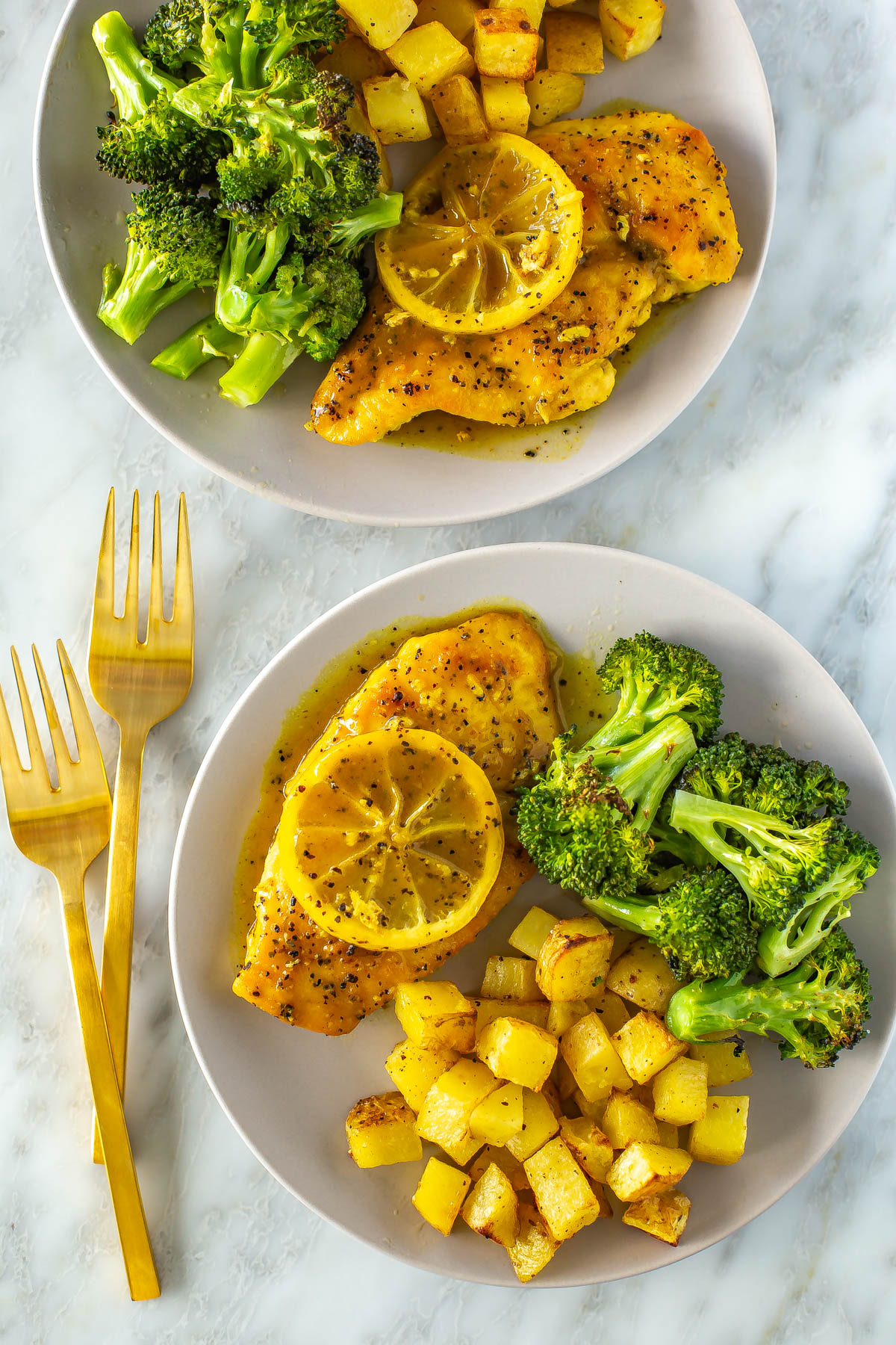 Two plates with lemon pepper chicken and a side of roasted potatoes and roasted broccoli.