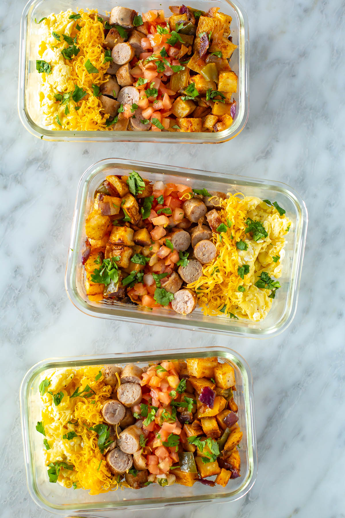 Three meal prep containers, each filled with roasted vegetables, scrambled eggs, and cooked sausage. 