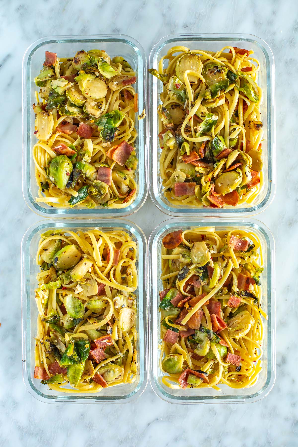 Four meal prep containers, each filled with a serving of creamy brussels sprouts pasta with bacon.