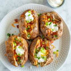 Four air fryer baked potatoes on a plate loaded with bacon, cheese, green onions, and sour cream.