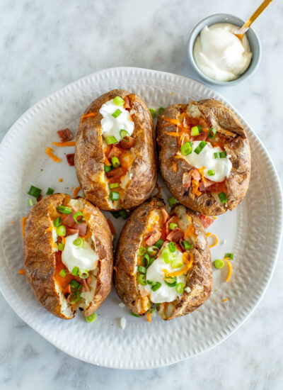 Four air fryer baked potatoes on a plate loaded with bacon, cheese, green onions, and sour cream.