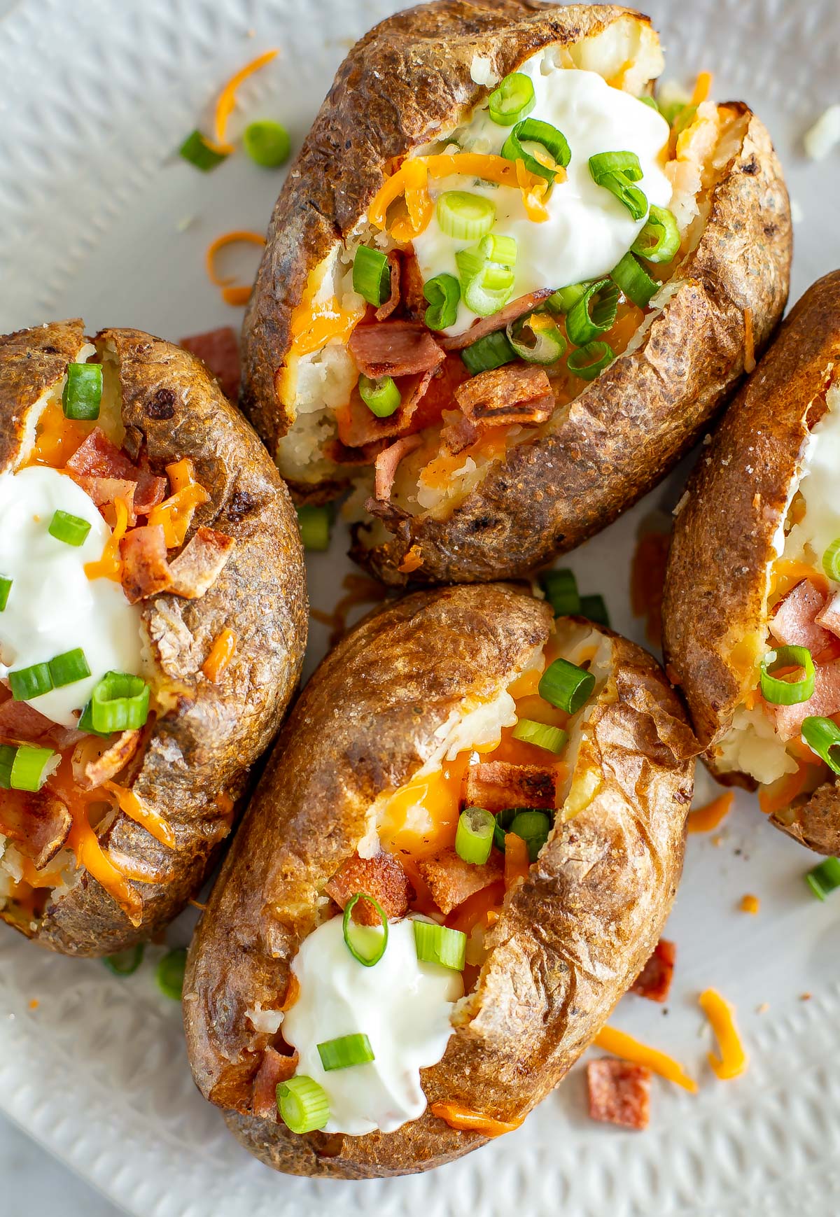 A close-up of four air fryer baked potatoes on a plate loaded with bacon, cheese, green onions, and sour cream.