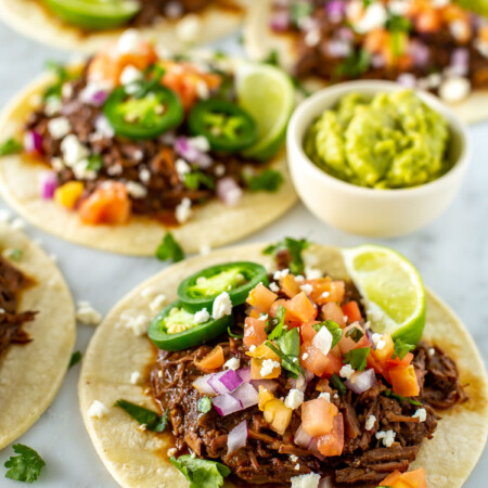 Four shredded beef tacos with a cup of guacamole on the side.