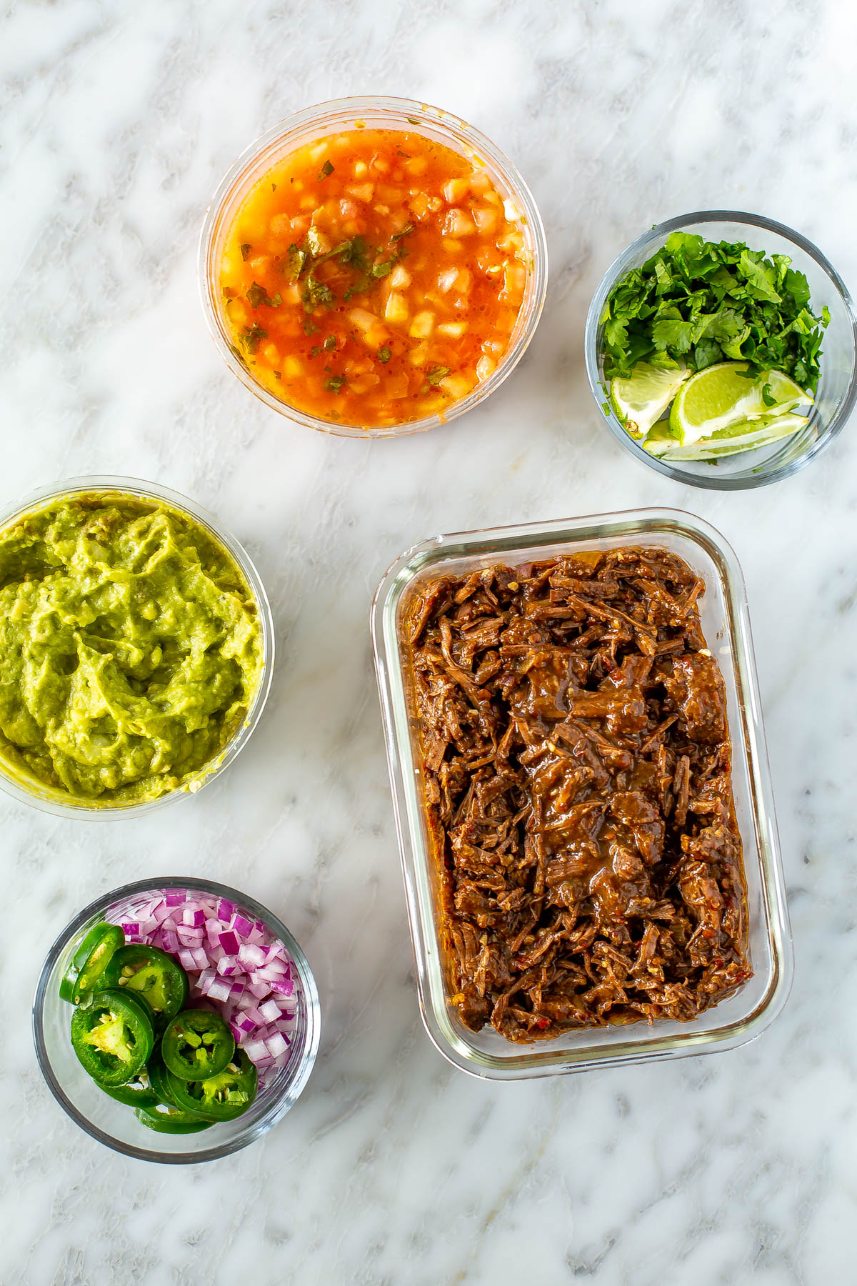 Meal prep containers filled with shredded beef, pico de gallo, guacamole, cilantro and lime wedges and jalapeno and red onion.