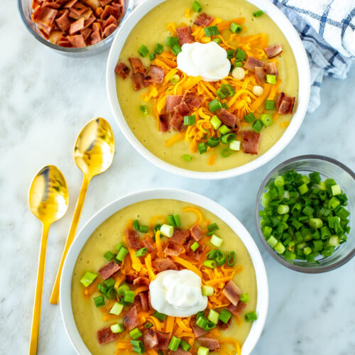Two bowls of crockpot potato soup topped with cheddar cheese, bacon, green onions and sour cream.