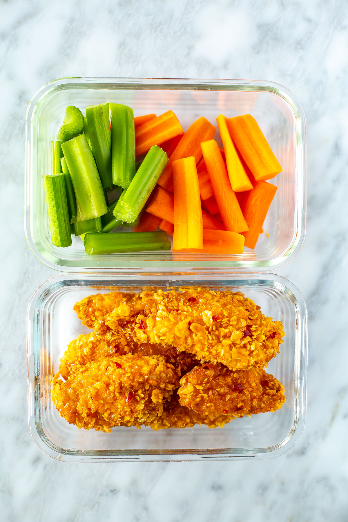 Two meal prep containers: one filled with air fryer hot honey chicken tenders and the other with chopped celery and carrots.