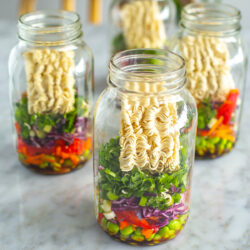 Four mason jars, each filled with instant noodles, vegetables and herbs.