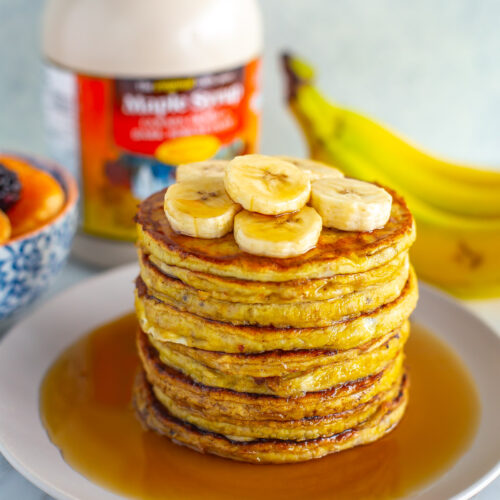 A stack of banana egg pancakes with extra banana slices and maple syrup on top.