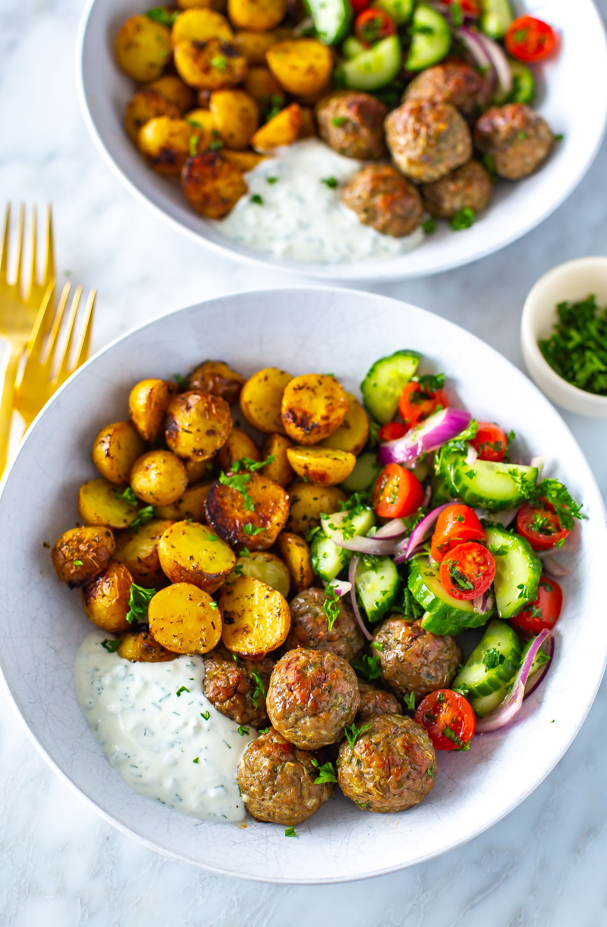 A close-up of a bowl of greek chicken meatballs with a side of potatoes, veggies and tzatziki. 
