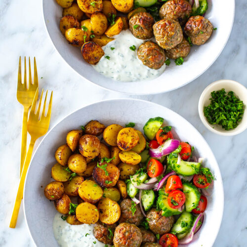 Two bowls of greek chicken meatballs with a side of potatoes, veggies and tzatziki.