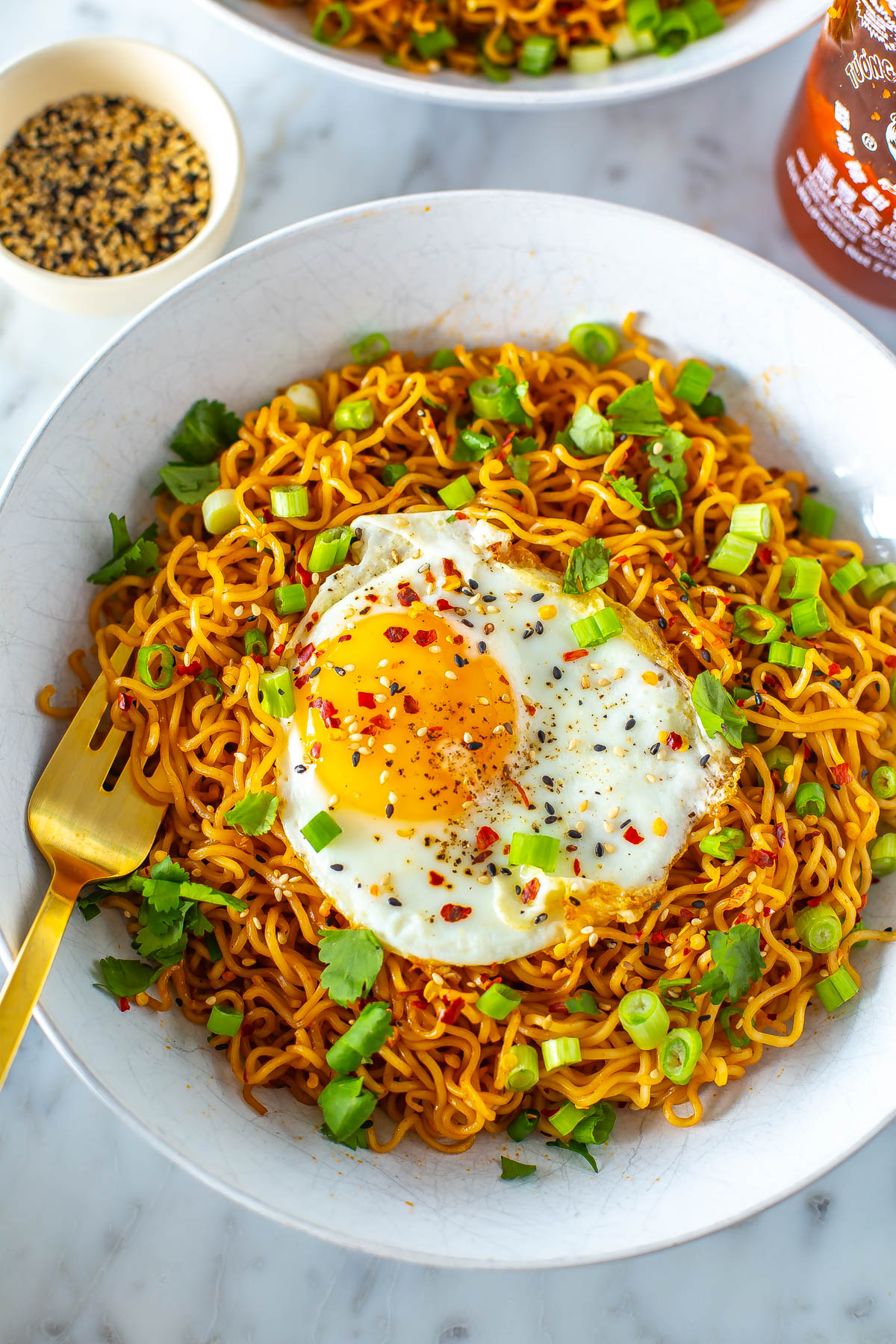 A close-up of a bowl of spicy noodles topped with a fried egg, green onions and cilantro with a fork in it.