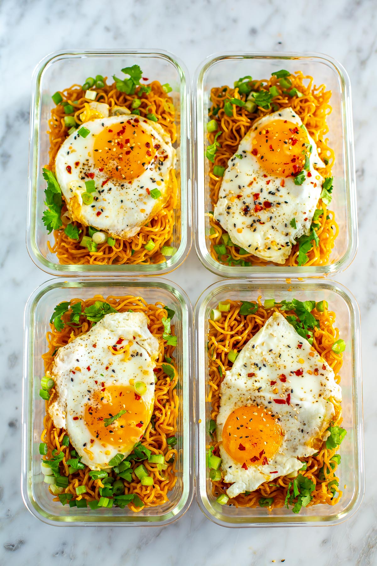Four meal prep containers, each with a serving of spicy noodles topped with a fried egg.