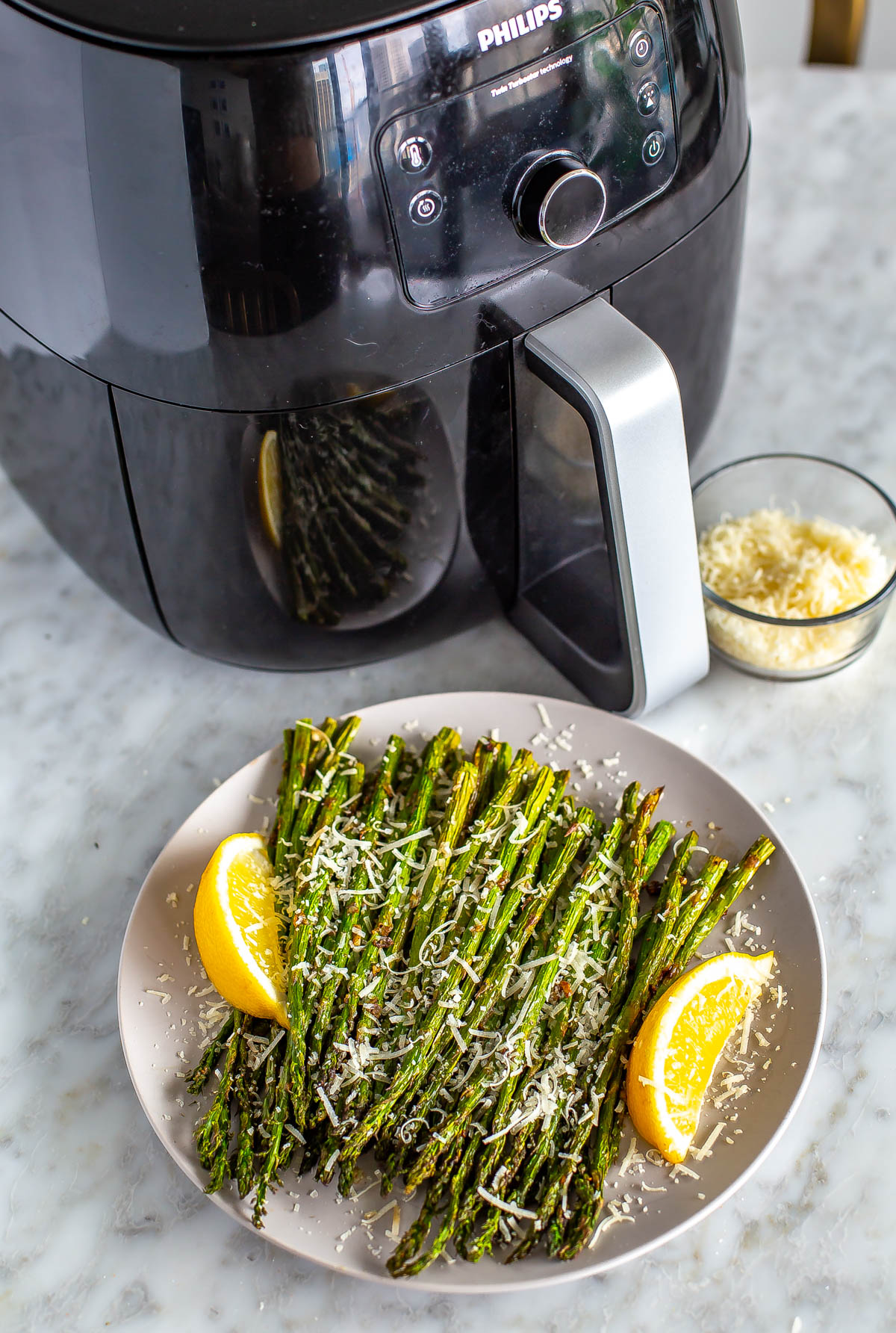 An air fryer in the background with a plate of garlic parm air fryer asparagus in the foreground.