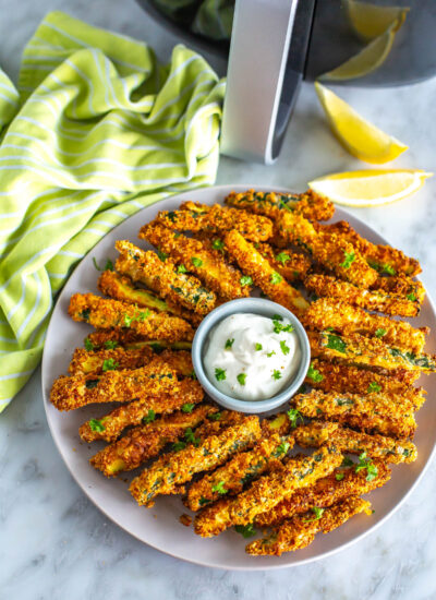 A plate of air fryer zucchini fries with garlic aioli in front of an air fryer.