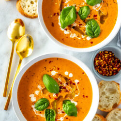 Two bowls of roasted tomato basil soup with baguette slices placed around it.