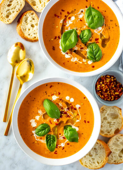 Two bowls of roasted tomato basil soup with baguette slices placed around it.
