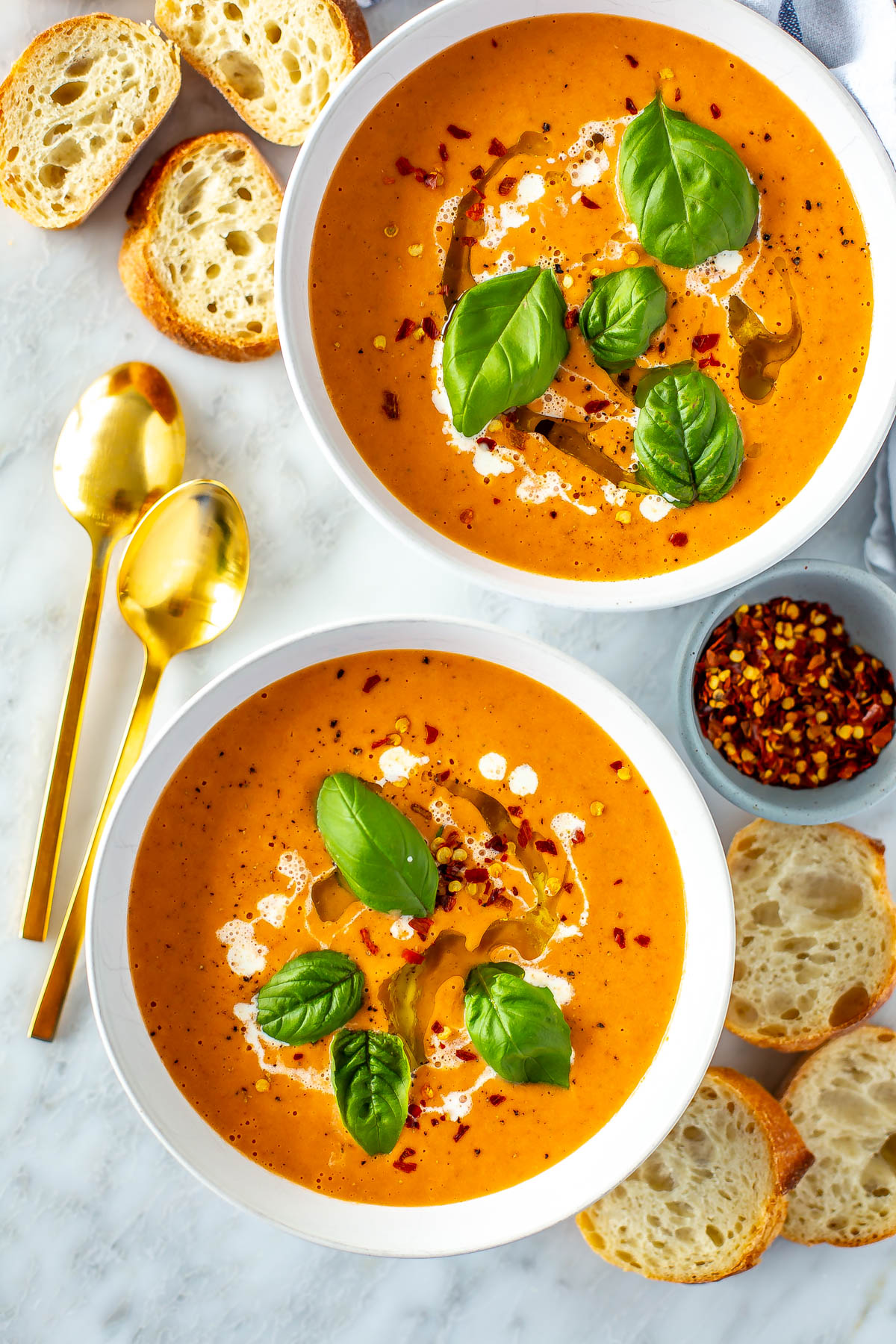Two bowls of roasted tomato basil soup with baguette slices placed around it. 