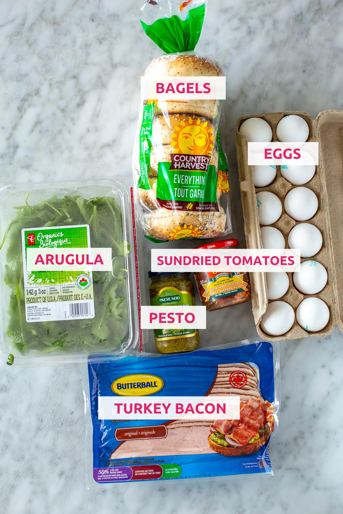 Ingredients for pesto bagel breakfast sandwiches: bagels, eggs, pesto, sundried tomatoes, arugula and turkey bacon.