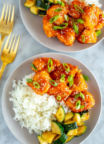 Two plates of firecracker chicken served alongside rice and zucchini.