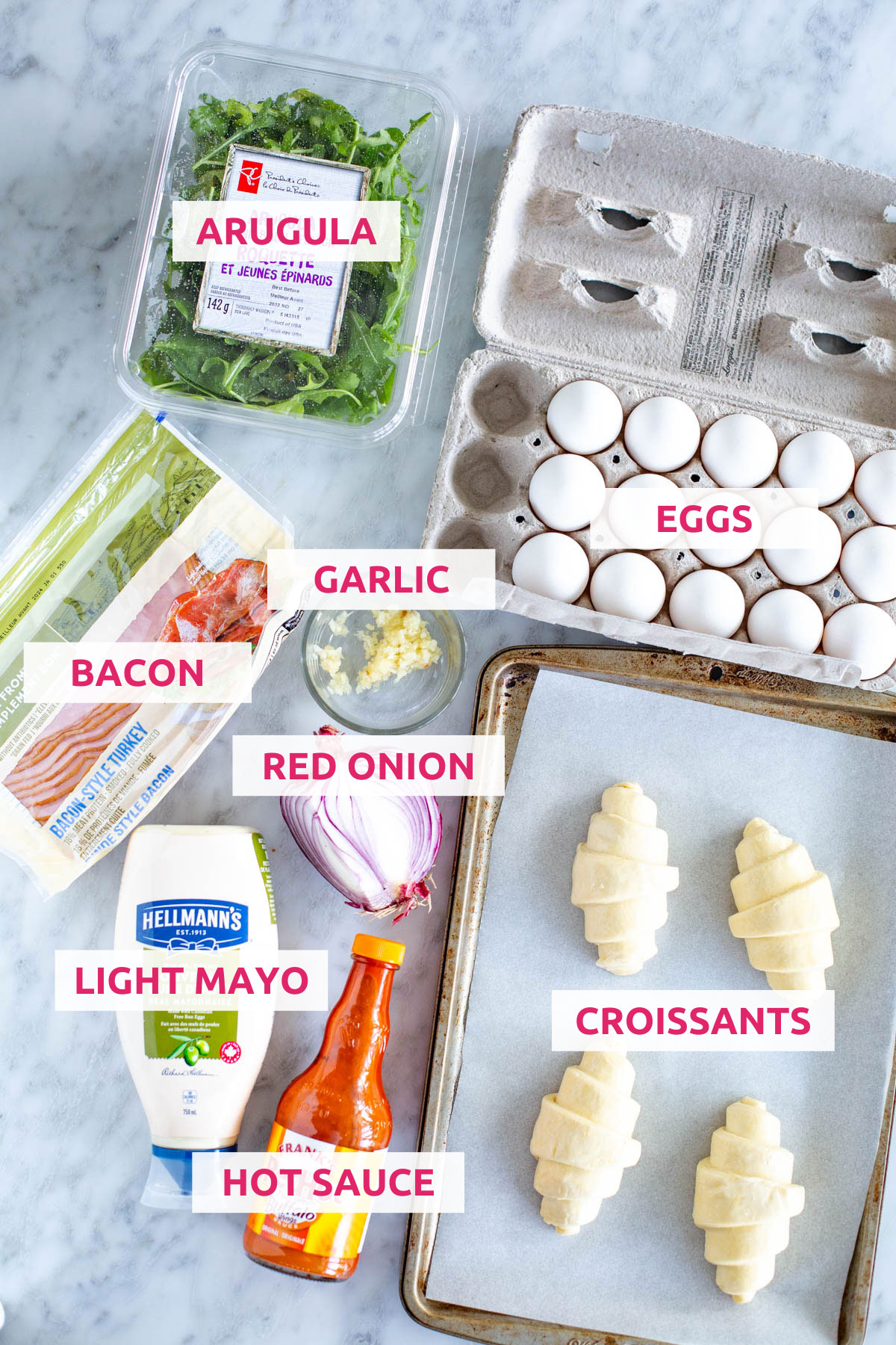 Ingredients for croissant breakfast sandwiches: croissants, eggs, arugula, bacon, garlic, hot sauce, light mayo and red onion.