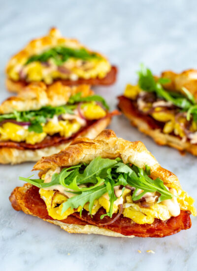 Four croissant breakfast sandwiches with bacon, scrambled eggs and arugula inside them.
