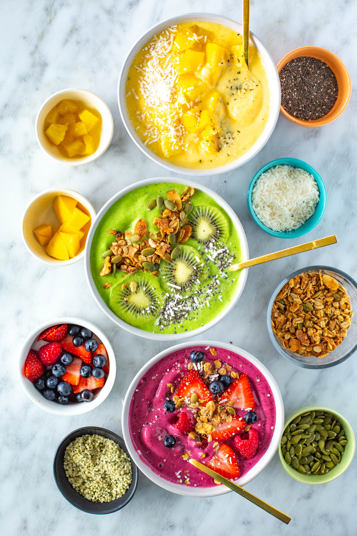 Three smoothie bowls, one yellow, one green and one pink, with various toppings.