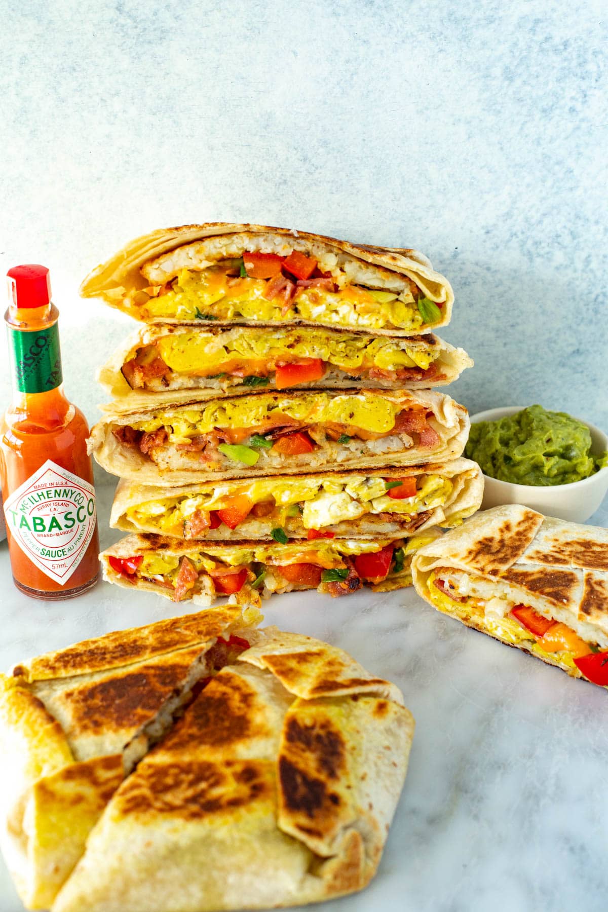 Cut open breakfast crunchwraps stacked together with a side of hot sauce and guacamole.